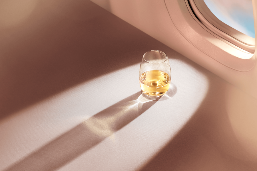 A stemless glass of white wine set in front of an aircraft window, with a long shadow extending from the glass. The sky's soft blush hue streams through the window and into the aircraft.